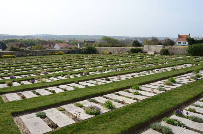 Vue d&rsquo;un cimeti&egrave;re militaire compos&eacute; de nombreuses tombes plates en pierre claire, dispos&eacute;es en rang&eacute;es r&eacute;guli&egrave;res sur une pelouse soigneusement entretenue. De petites plantes et des fleurs poussent entre certaines tombes. Le cimeti&egrave;re est entour&eacute; d&rsquo;un mur de pierre ; au fond, un monument comm&eacute;moratif en pierre claire se dresse devant le mur. Au-del&agrave;, on aper&ccedil;oit quelques maisons et un paysage vallonn&eacute; sous un ciel clair.