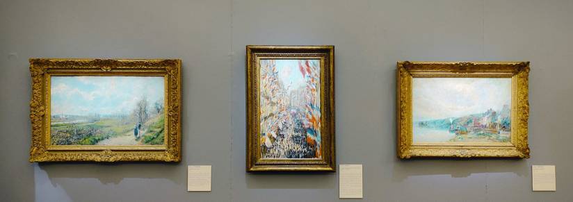 PHOTO 1 : Trois tableaux sont disposés sur un mur gris. La peinture centrale est au format portrait et représente une fête nationale, c’est le tableau « La Rue Montorgueil, à Paris. Fête du 30 juin 1978 » par Claude Monet.  Les deux peintures sur les côtés sont en format paysage et présentent des paysages impressionnistes.