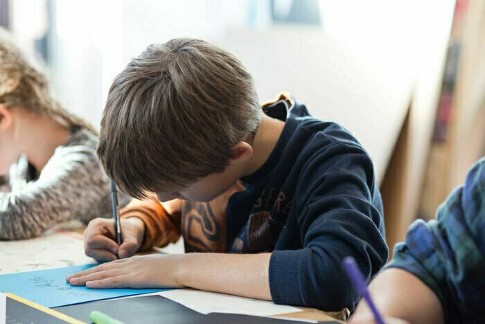 PHOTO 13 : Un jeune garçon est assis à une table et écrit ou dessine sur une feuille en papier. On aperçoit également d’autres enfants en arrière-plan.