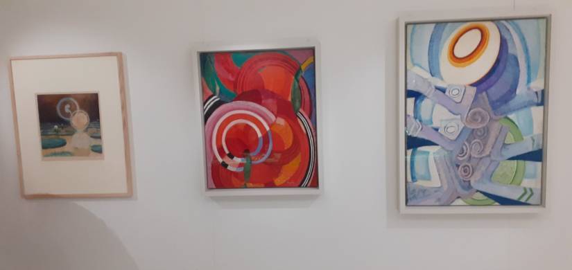 PHOTO 3 : Trois peintures sont accrochées à un mur blanc. La peinture à gauche représente une scène abstraite avec des couleurs douces et des formes circulaires. L'œuvre du milieu est plus vive, avec des cercles concentriques rouges, roses, et oranges. La peinture à droite utilise des teintes de bleu et de violet, avec des motifs circulaires.