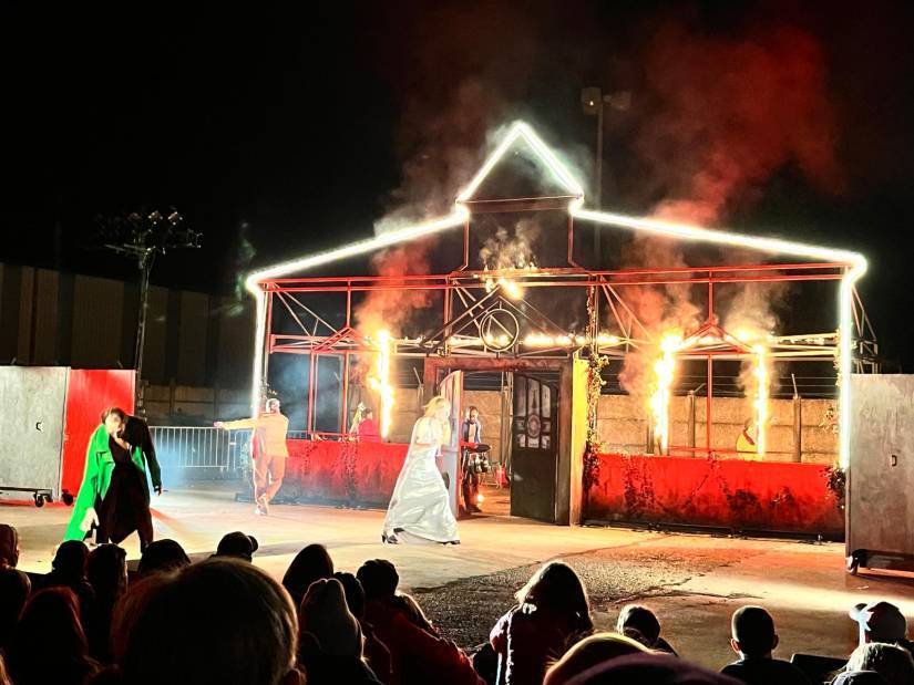 PHOTO 2 : Une performance de spectacle est réalisée en plein air, de nuit. Une structure en forme de maison, est illuminée par des néons blancs et entourée de flammes. Au premier plan, plusieurs artistes sont en costume, dont une personne en robe argentée et une autre en manteau vert vif. La foule assise regarde la scène