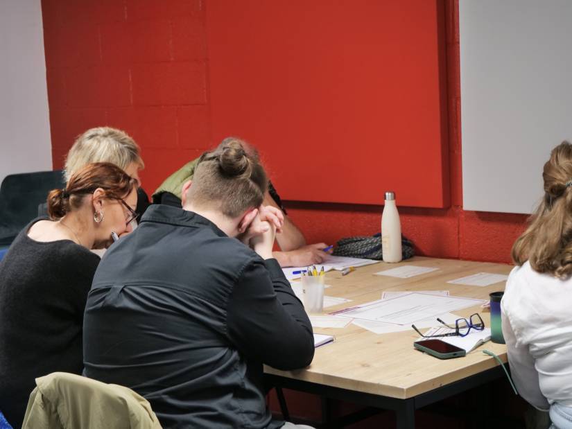Quatre personnes sont attablées de dos autour d'un exercice, et semblent concentrées.