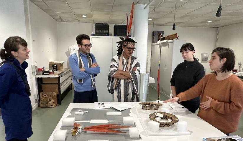 Un repr&eacute;sentant d'une communaut&eacute; autochtone amazonienne, en tenue traditionnelle, assiste &agrave; une pr&eacute;sentation d'ornements de t&ecirc;te issus de sa culture, pr&eacute;sent&eacute;s sur une table devant lui par plusieurs professionnels de l'Ecole sup&eacute;rieure d'art d'Avignon.