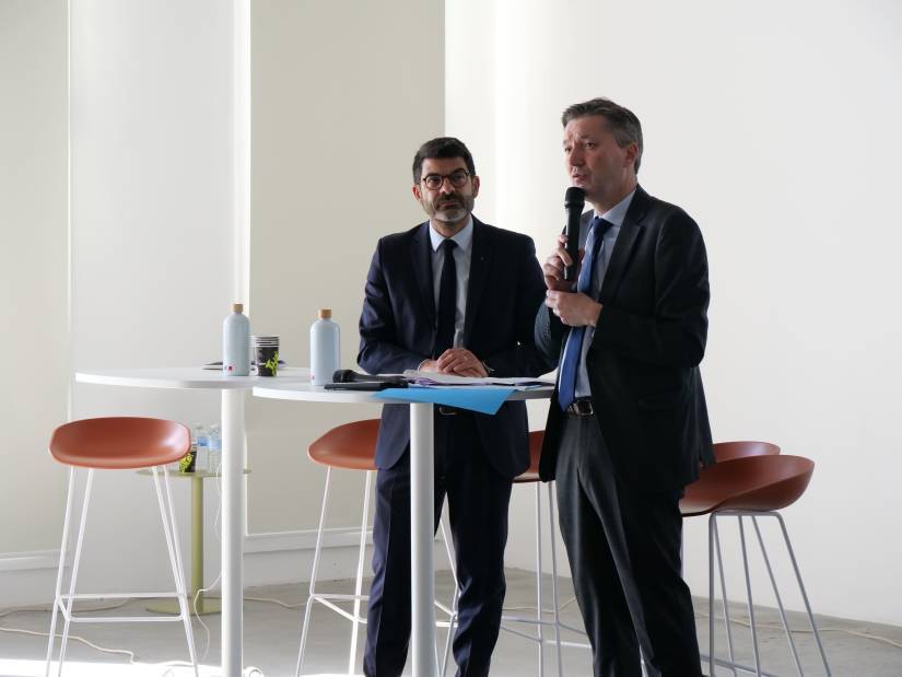 Le préfet Bertrand Gaume (à droite) prend la parole en tenant un micro, debout à côté de Hilaire Multon, directeur régional des affaires culturelles des Hauts-de-France, à sa gauche. Les deux hommes, en costume sombre, se tiennent près d’une table haute ronde sur laquelle sont posés des documents, deux gourdes ornées du drapeau français et des gobelets. La scène se déroule dans une salle lumineuse, sobrement aménagée avec des chaises hautes et un mur blanc en arrière-plan. L’atmosphère est formelle, probablement dans le cadre d’un discours ou d’une conclusion de séminaire ou de réunion institutionnelle.