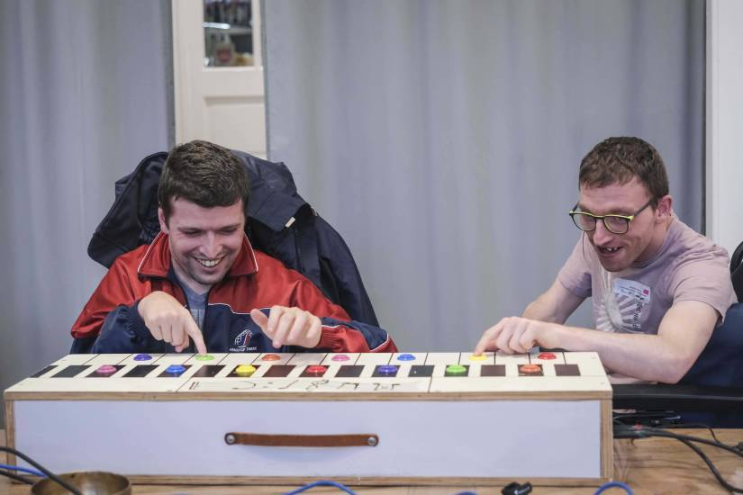 Deux jeunes adultes assis à une table jouent sur un grand instrument de musique fabriqué à partir de touches colorées et de larges boutons. Tous deux sourient en appuyant sur les touches, concentrés et manifestement heureux de produire des sons.