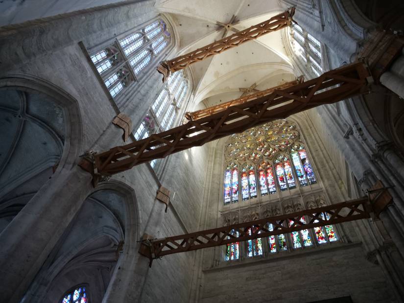 De grandes poutres en poids traverses de part et d'autre la voûte de la cathédrale, dans une vue contre-plongée et sous la lumière des vitraux.