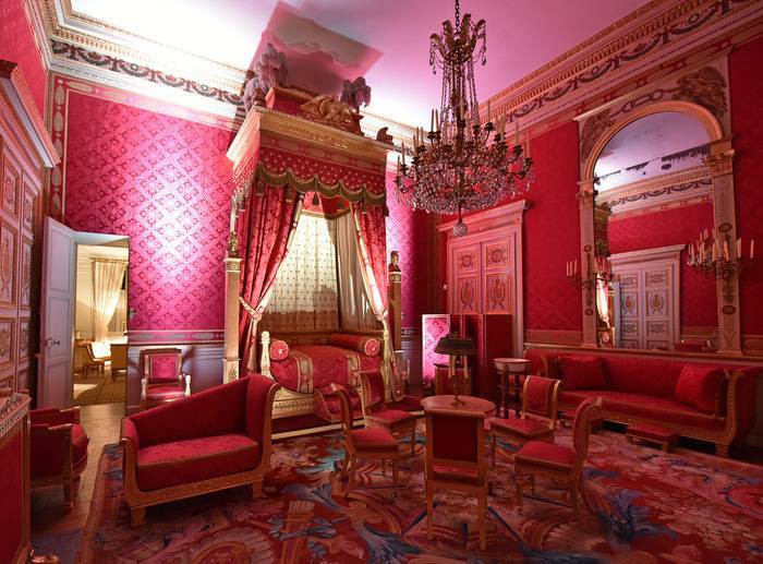 PHOTO 5 : Chambre de château, richement décorée et entourée de murs recouverts de tapisseries rouges. Au niveau des meubles, on retrouve un lit à baldaquin et des fauteuils dans les tons rouges également.