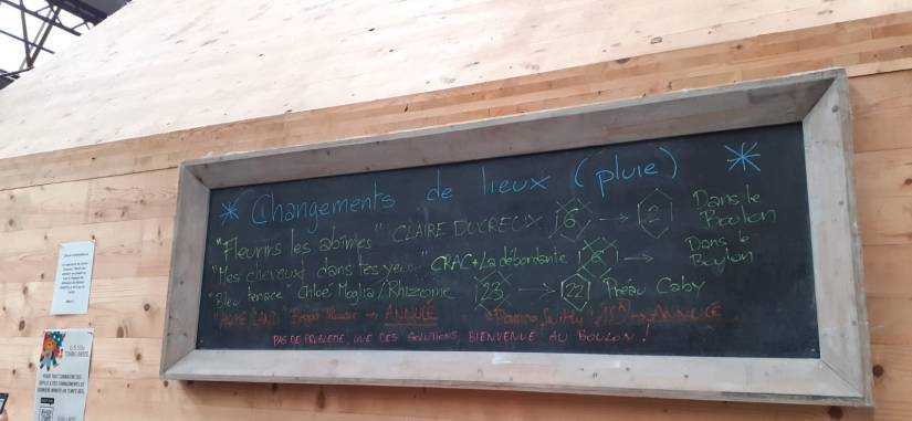 PHOTO 1 : Une ardoise est accrochée sur un mur en bois, avec des inscriptions à la craie. Le tableau annonce des changements de lieux en raison de la pluie, pour plusieurs spectacles. On peut lire des titres et des flèches et des notes indiquent les nouveaux lieux.