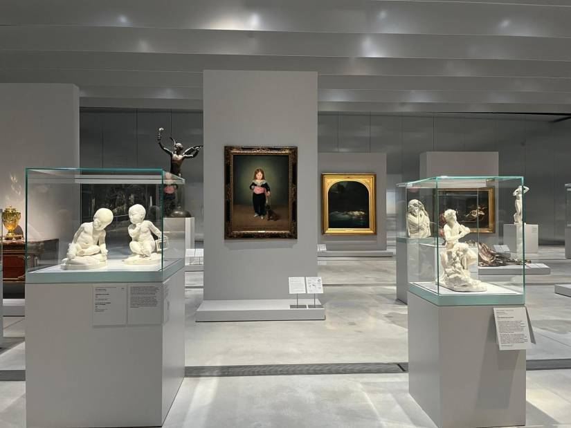 Vue d'une salle d'exposition, présentant des œuvres variées. Au centre, un tableau de Francisco de Goya représentant un jeune enfant vêtu de noir, tenant des objets dans ses mains, entouré de sculptures sous vitrines. À gauche, deux bustes d’enfants en marbre blanc se font face dans une vitrine. À droite, plusieurs sculptures, dont une représentant un personnage en mouvement. L’ensemble est disposé dans une ambiance lumineuse et épurée, mettant en valeur les œuvres exposées.