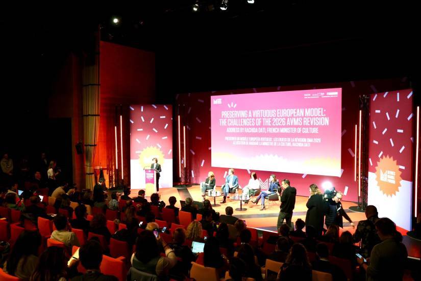 Une conférence dans le cadre du Séries Mania Forum, avec un public nombreux installé dans des fauteuils orange et rouges. Sur scène, Rachida Dati, ministre de la Culture, s'exprime depuis un pupitre, tandis qu’un panneau lumineux derrière elle affiche le titre de son discours en anglais et en français. Cinq intervenants sont assis à sa droite. L’ambiance est dynamique avec des lumières rouges et un décor graphique.