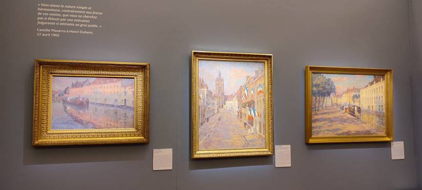 PHOTO 3 : Trois peintures impressionnistes sont accrochées sur un mur gris. Les peintures représentent des scènes urbaines avec des bâtiments, des rues et des canaux. Chaque tableau est encadré par un cadre doré.