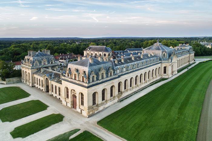 PHOTO 2 : Vue aérienne du domaine de Chantilly. Le bâtiment est caractérisé par une architecture classique. À l'arrière-plan, une forêt dense s'étend, tandis qu'au premier plan, des allées de gravier et des pelouses encadrent le château.
