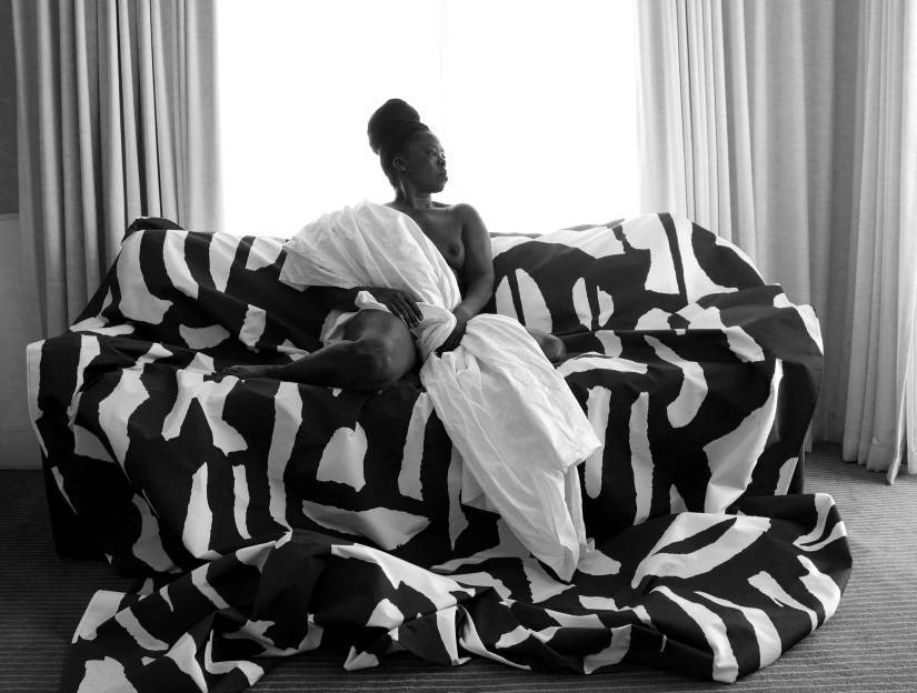 photo en noir et blanc d'une femme noire assise dans un canapé dans un décor luxueux. un drap noir et blanc est posé sur le canapé et la femme est enroulée dans un drap blanc et regarde sur le côté