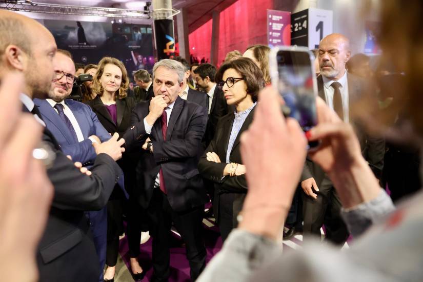 Une scène d'échange au Séries Mania Forum, où plusieurs personnes en tenue formelle sont rassemblées sous un éclairage tamisé aux teintes rouges et violettes. Au centre, un homme en costume sombre, cravate bordeaux, main sur le menton, semble écouter attentivement : on reconnait Xavier Bertrand, président de la région Hauts-de-France. À ses côtés, une femme en costume noir croise les bras avec une expression concentrée : il s'agit de Rachida Dati,, ministre de la Culture. D'autres participants, en arrière-plan, observent ou échangent. Au premier plan, une personne prend une photo avec un smartphone, floutant une partie de l'image. Des affiches et panneaux aux couleurs du festival Série Mania sont visibles en fond, suggérant un contexte lié à cet événement culturel.