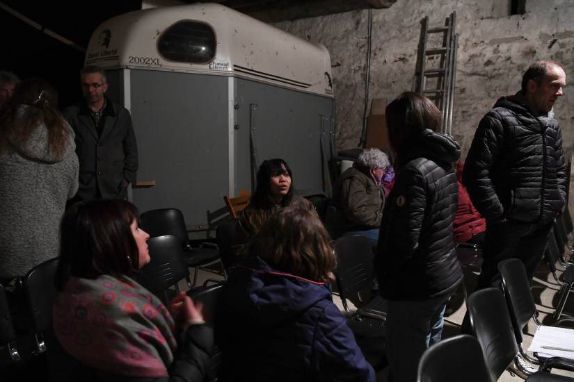 PHOTO 1 : Des individus se lèvent de leurs chaises. On peut voir en arrière-plan un van qui transporte des chevaux, on comprend bien qu’il s’agit d’une salle qui n’a pas vocation à être une salle de projection.
