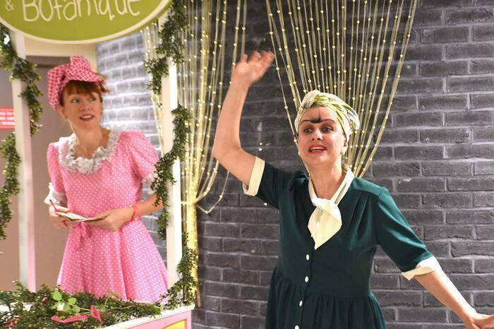 PHOTO 15 : Deux femmes en costume participent à une scène de spectacle. La femme à gauche porte une robe rose à pois blancs avec un col bouffant. Elle se tient derrière un comptoir. La femme à droite porte une robe verte foncée avec des manches et un col blanc.