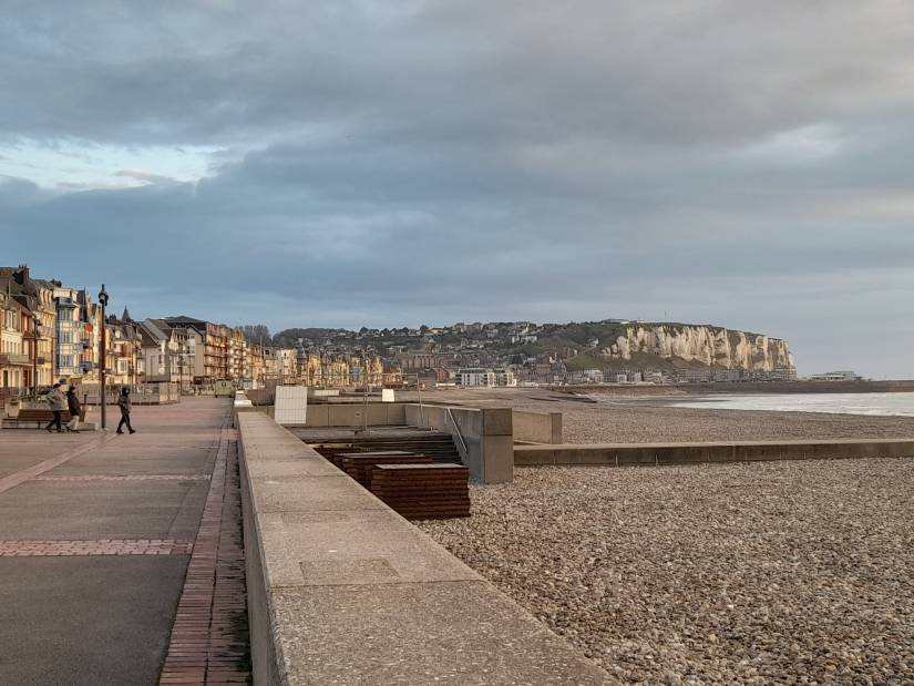Vue d'une promenade en bord de mer, bordée de maisons colorées à l'architecture balnéaire. À droite, une plage de galets s'étend vers l'horizon, dominée par des falaises blanches caractéristiques. Le ciel nuageux laisse passer une lumière douce, et quelques passants profitent de la promenade dans une atmosphère paisible.