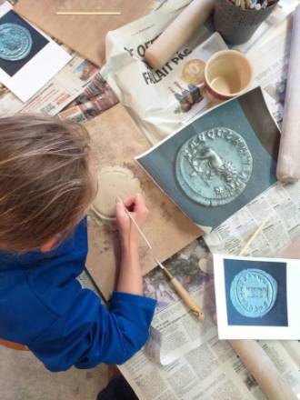 PHOTO 7 : Une personne travaille sur une plaque d'argile, entourée d'outils de modelage et de feuilles de papier avec des images de médaillons anciens.