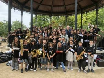 PHOTO 2 : Un chœur d’enfants pose sous un kiosque à musique en plein air. Ils tiennent différents instruments de musique, tels que des saxophones, des trompettes, des cors, et des clarinettes.