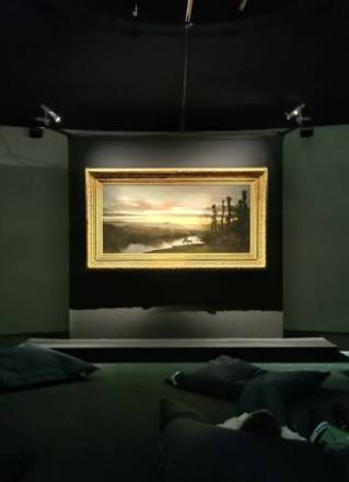 PHOTO 5 : Une salle sombre met en valeur un tableau accroché au mur. Le tableau représente un paysage. Devant le tableau, des coussins sont disposés au sol, permettant aux visiteurs de s'asseoir ou de s'allonger pour admirer l'œuvre d'art.