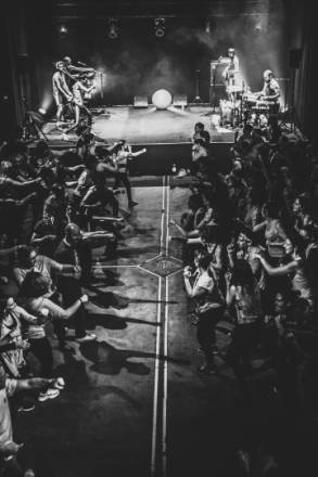 Photo 6 : Cette photo en noir et blanc représente une scène de concert. À l'arrière-plan, des musiciens jouent sur scène avec leurs instruments. Au premier plan, une foule dense et enthousiaste danse.
