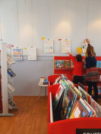 photo dans une biblioth&egrave;que avec des livres au premier plan et deux personnes accrochant des dessins au mur &agrave; l'arri&egrave;re plan