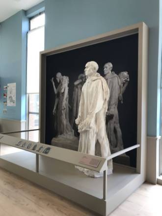 Vue d’une salle d’exposition du musée des beaux-arts de Calais, présentant un moulage en plâtre d’un des personnages du groupe sculpté Les Bourgeois de Calais d’Auguste Rodin. La figure représentée au premier plan est celle d’Eustache de Saint-Pierre, tête nue et mains jointes tenant la clé de la ville, vêtu d’un long manteau. Elle se détache devant une photographie en noir et blanc représentant le groupe complet des six bourgeois en arrière-plan. À l’avant, un pupitre incliné affiche des portraits et un cartel explicatif. Les murs de la salle sont peints en bleu clair et un grand vitrage latéral laisse passer la lumière naturelle.