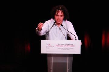 photo d'un jeune homme derri&egrave;re un pupitre en train de parler en tendant le bras