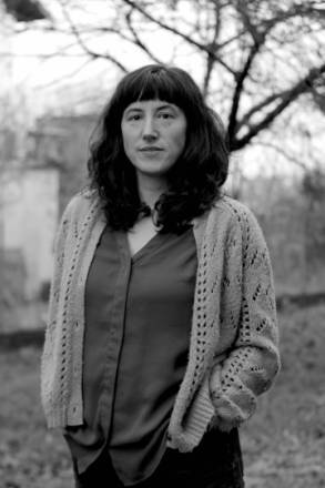 portrait en noir et blanc d'aurore petit en extérieur, les mains dans les poches, cheveux bruns long avec une frange avec un arbre en arrière plan