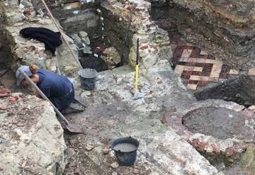 Une arch&eacute;ologue &agrave; genoux d&eacute;gage &agrave; la truelle un ensemble constitu&eacute; de deux petites pi&egrave;ces carrel&eacute;es s&eacute;par&eacute;es par une entr&eacute;e avec escalier. Devant, un puits attend d'&ecirc;tre fouill&eacute;.