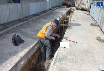 Un arch&eacute;ologue portant un gilet de s&eacute;curit&eacute; est debout dans une tranch&eacute;e. Cette tranch&eacute;e, profonde de 1 m environ, est ouverte dans la rue. L'arch&eacute;ologue prend des mesures qu'il reporte sur le dessin pos&eacute; &agrave; c&ocirc;t&eacute; de lui sur le bitume.