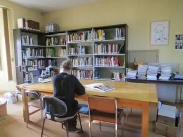Assise &agrave; la grande table de lecture, une arch&eacute;ologue feuillette des livres. Au fond, un rayonnage charg&eacute; d'ouvrages et un bureau o&ugrave; sont empil&eacute;s des rapports de fouilles.