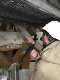 Un homme avec un casque de sécurité desserre un boulon sur une charpente en bois et en métal de grande dimension.