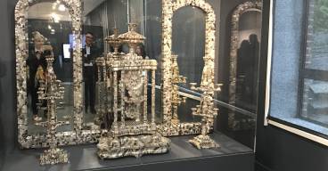 Vue d’ensemble d’une vitrine d’exposition du musée de la Nacre et de la Tabletterie à Méru. Au centre, un monumental autel en nacre finement sculptée, composé d’éléments architecturaux et ornementaux, avec colonnes, baldaquin et candélabres. L’ensemble est présenté dans une vitrine vitrée avec des miroirs en fond, amplifiant l’effet visuel. Une femme prenant une photo est visible, reflétée dans le miroir central. La scène est éclairée par des spots au plafond et baignée d’une lumière tamisée.