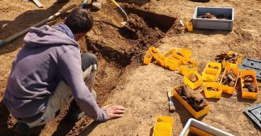 Un archéologue est accroupi dans un petit fossés en V. Il fouille à la truelle et dépose ses découvertes dans de petites boîtes en plastique jaune.