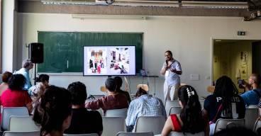 photo d'un homme debout avec un micro à la main, en train de présenter quelque chose à un public assis, de dos