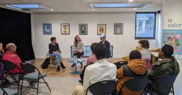 photo avec une petite conférence dans une bibliothèque avec du public de dos et trois intervenants de face assis