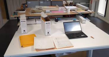 Sur des tables sont étalés des plans, des boîtes d'archives et des fiches d'enregistrement et un ordinateur ouvert. Sous la table, un grand cartons dans lesquels sont jetés tous les brouillons.