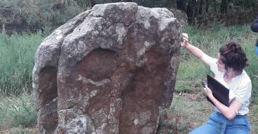 Une jeune femme observe un menhir en vue d'établir un constat d'état.