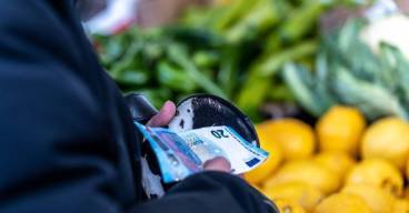 photo d'une personne tenant son porte monnaie avec des billets devant un stand de fruits et légumes