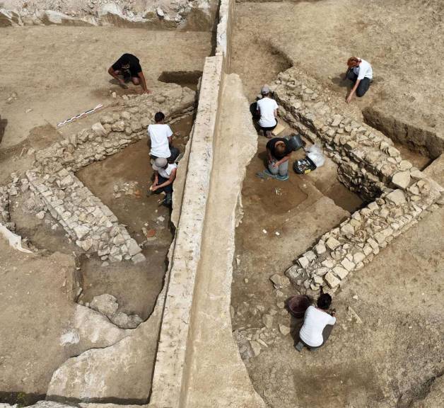 Vu du ciel : un petit bâtiment carré arasé est traversé par une berme de fouille. De chaque côté, des fouilleurs sont à genoux pour fouiller finement les niveaux anciens.au travail.