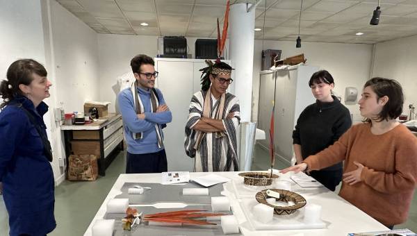 Un représentant d'une communauté autochtone amazonienne, en tenue traditionnelle, assiste à une présentation d'ornements de tête issus de sa culture, présentés sur une table devant lui par plusieurs professionnels de l'Ecole supérieure d'art d'Avignon.