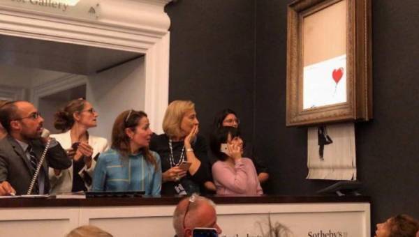 Dans une salle de vente aux enchères, des enchérisseurs regardent, effarés, un tableau encadré de l'artiste Banksy qui s'autodétruit.