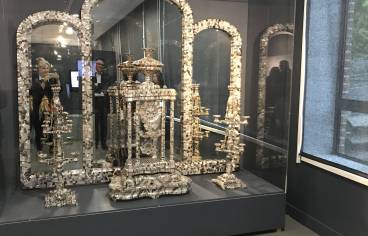 Vue d’ensemble d’une vitrine d’exposition du musée de la Nacre et de la Tabletterie à Méru. Au centre, un monumental autel en nacre finement sculptée, composé d’éléments architecturaux et ornementaux, avec colonnes, baldaquin et candélabres. L’ensemble est présenté dans une vitrine vitrée avec des miroirs en fond, amplifiant l’effet visuel. Une femme prenant une photo est visible, reflétée dans le miroir central. La scène est éclairée par des spots au plafond et baignée d’une lumière tamisée.
