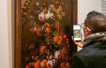 Photo de couverture : Un individu prend une photo d'une peinture avec son téléphone portable. La peinture représente une nature morte avec un bouquet de fleurs et des fruits.