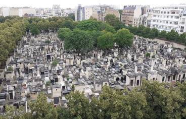 Vue aérienne du cimetière du Montparnasse, dans le 14e arrondissement de Paris, aux multiples tombes et caveaux en pierres, serrés les uns contre les autres. Le cimetière est bordé d'arbres aux feuillages denses, très verts et accueille un bosquet tout aussi vert en son centre. Autour du cimetière, des immeubles résidentiels et des bâtiments urbains sont visibles, formant une bordure claire entre la ville et le cimetière. La perspective donne une impression de densité et d'ordre dans l'aménagement du lieu.