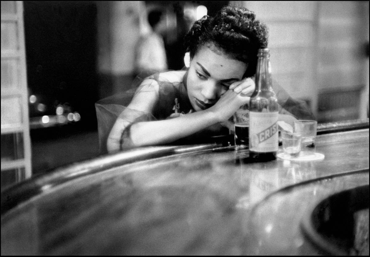 photo en noir et blanc d'Eve Arnold d'une femme accoudé à un bar avec une bouteille et un verre et qui regarde sur le côté, l'air songeur
