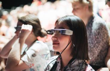 photo avec au premier plan une jeune femme portant des lunettes reliées à un fil, en train de regarder un spectacle. Au second plan, deux autres personnes dont une a aussi ces lunettes