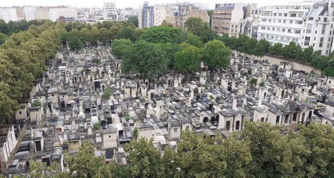 Vue aérienne du cimetière du Montparnasse, dans le 14e arrondissement de Paris, aux multiples tombes et caveaux en pierres, serrés les uns contre les autres. Le cimetière est bordé d'arbres aux feuillages denses, très verts et accueille un bosquet tout aussi vert en son centre. Autour du cimetière, des immeubles résidentiels et des bâtiments urbains sont visibles, formant une bordure claire entre la ville et le cimetière. La perspective donne une impression de densité et d'ordre dans l'aménagement du lieu.