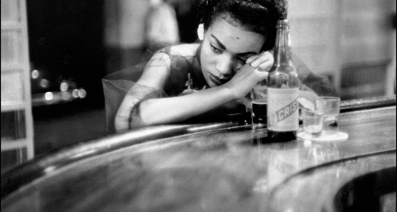 photo en noir et blanc d'Eve Arnold d'une femme accoudé à un bar avec une bouteille et un verre et qui regarde sur le côté, l'air songeur